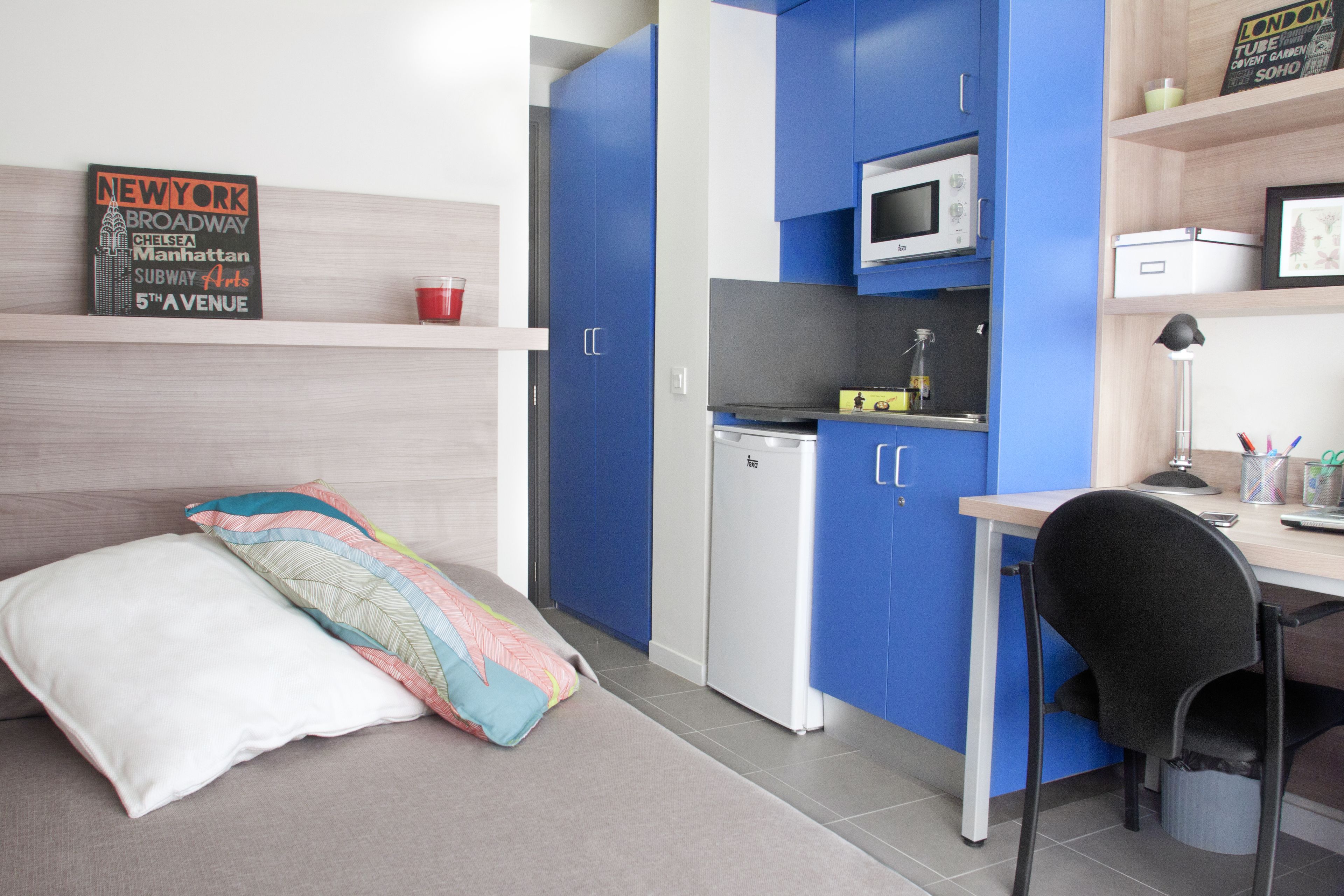 Una kitchenette compacta con microondas, un refrigerador pequeño y un lavamanos. Se ve una cama con un almohadón de colores y un almohadón blanco. Una estantería muestra un letrero y una taza roja.