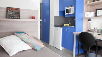Una kitchenette compacta con microondas, un refrigerador pequeño y un lavamanos. Se ve una cama con un almohadón de colores y un almohadón blanco. Una estantería muestra un letrero y una taza roja.