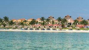 A beachfront hotel with thatched-roof buildings, palm trees, and a row of beachside chairs. - El Beso Adults Only at Ocean Coral & Turquesa - All inclusive (Puerto Morelos)