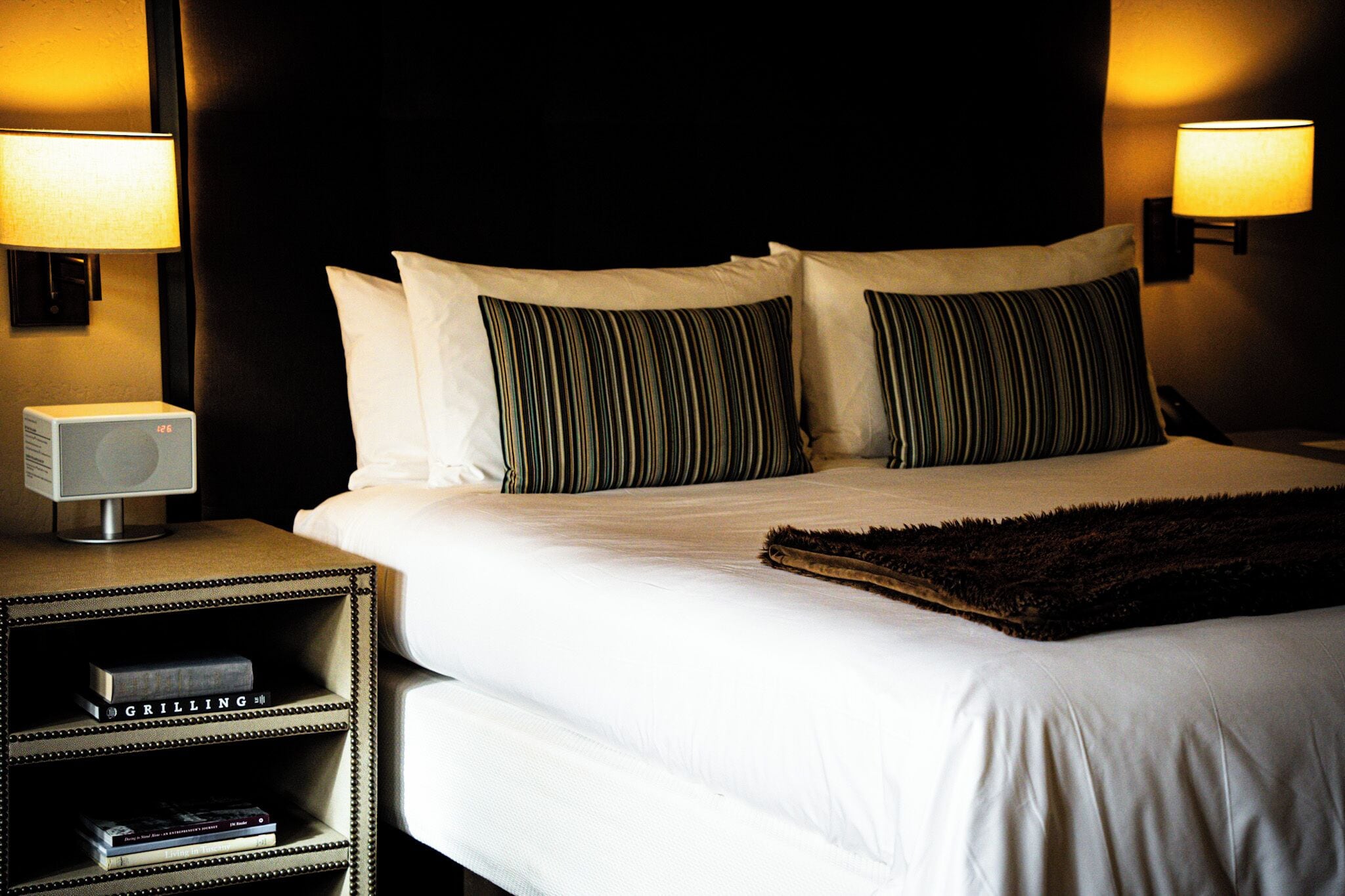 A neatly made bed with striped pillows, a dark throw, and two bedside lamps. There is a small shelf with books and a digital clock.
