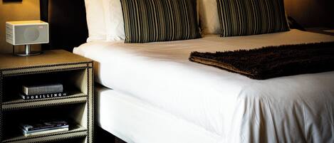 A neatly made bed with striped pillows, a dark throw, and two bedside lamps. There is a small shelf with books and a digital clock.
