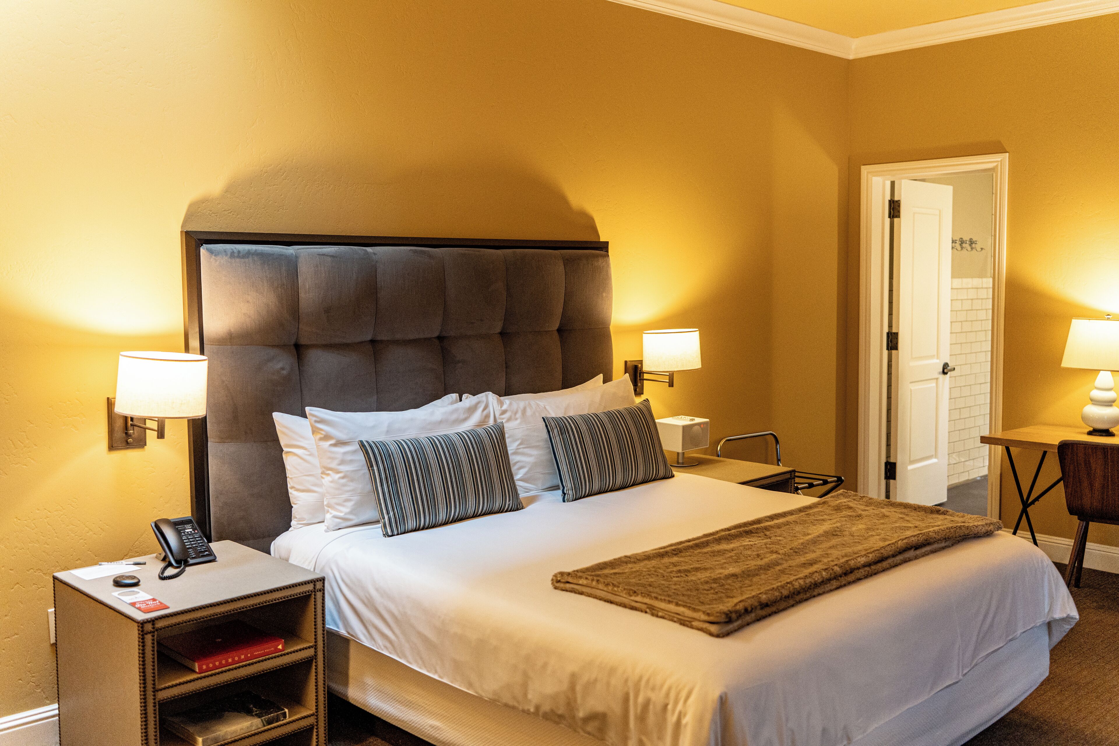 A hotel room with a large bed, two bedside lamps, a bedside table with a lamp, a telephone, and a small shelf with books.