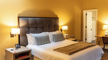A hotel room with a large bed, two bedside lamps, a bedside table with a lamp, a telephone, and a small shelf with books.