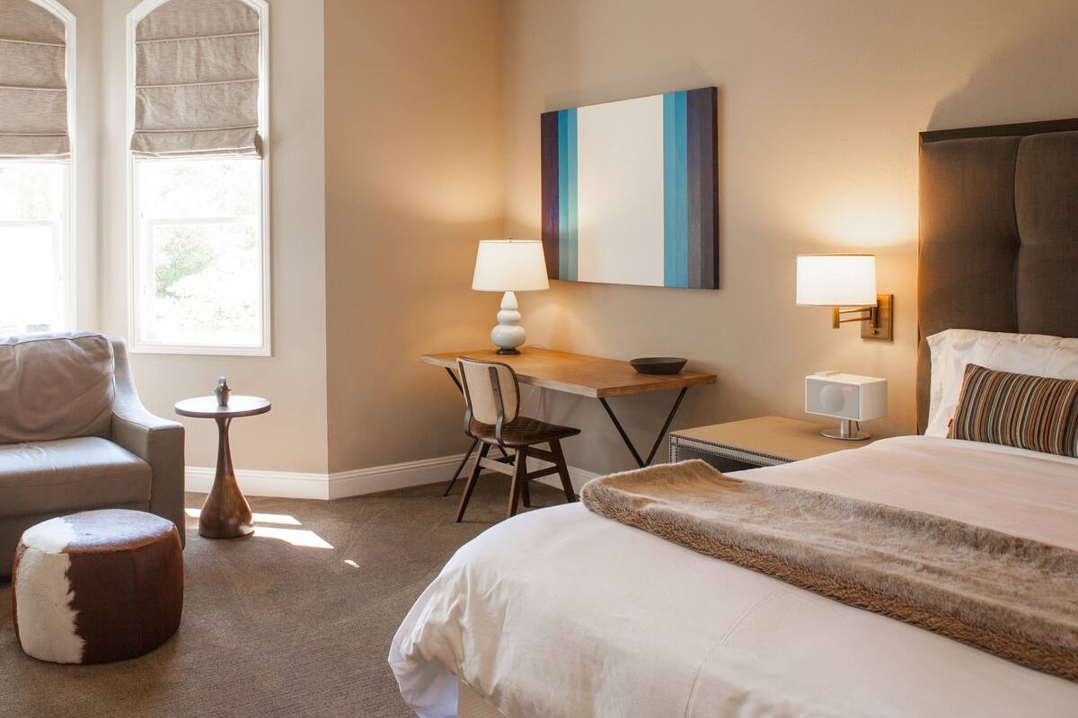 A hotel room with a bed, a desk with a chair, a small table, and a lamp.