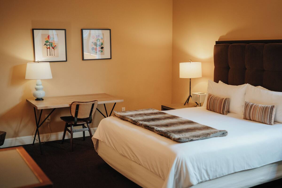 A hotel room with a bed, two bedside lamps, a desk with a chair, and two framed pictures on the wall.