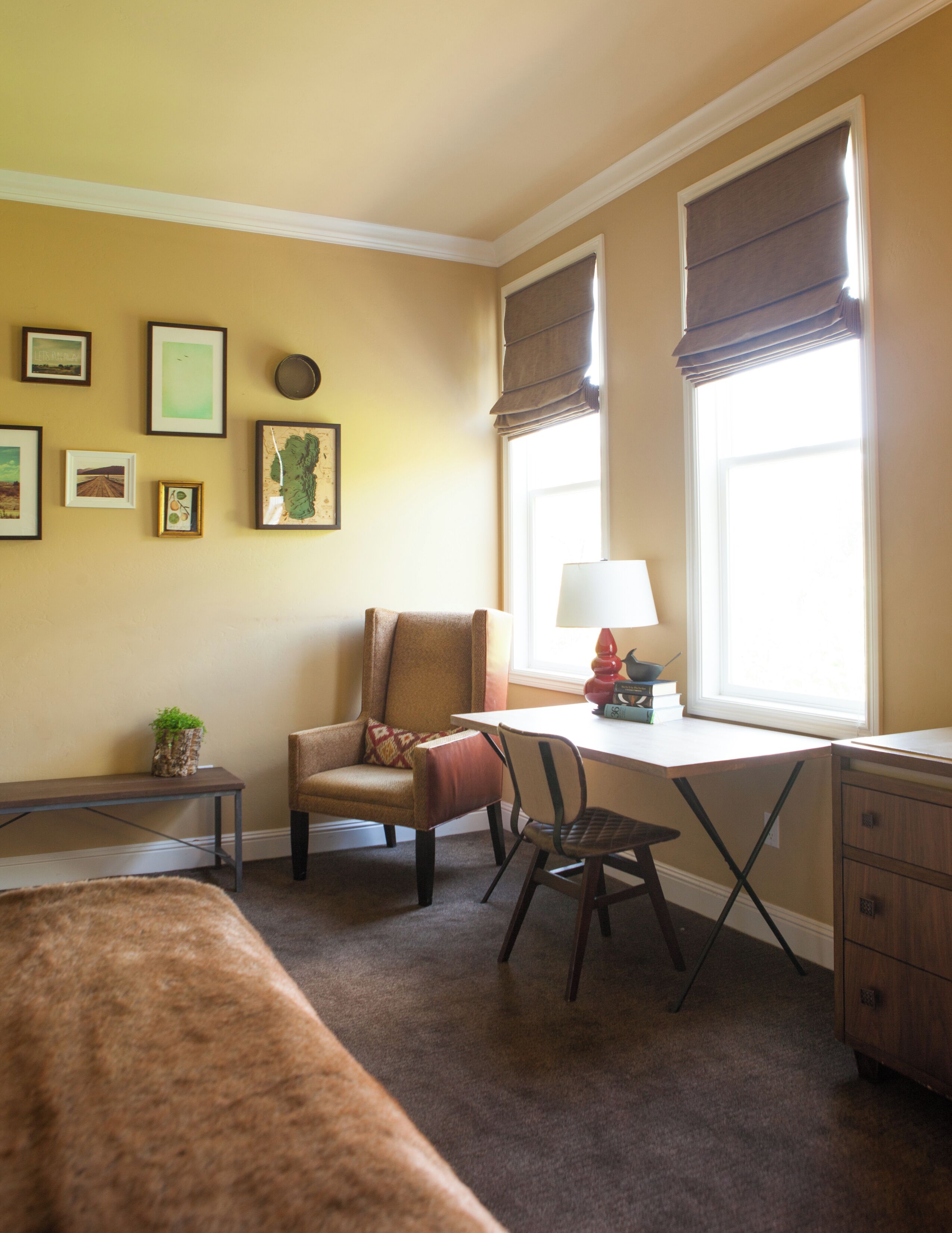 Una habitación con escritorio, silla y cama. Hay cuadros en la pared, una lámpara en el escritorio y una ventana con persianas.
