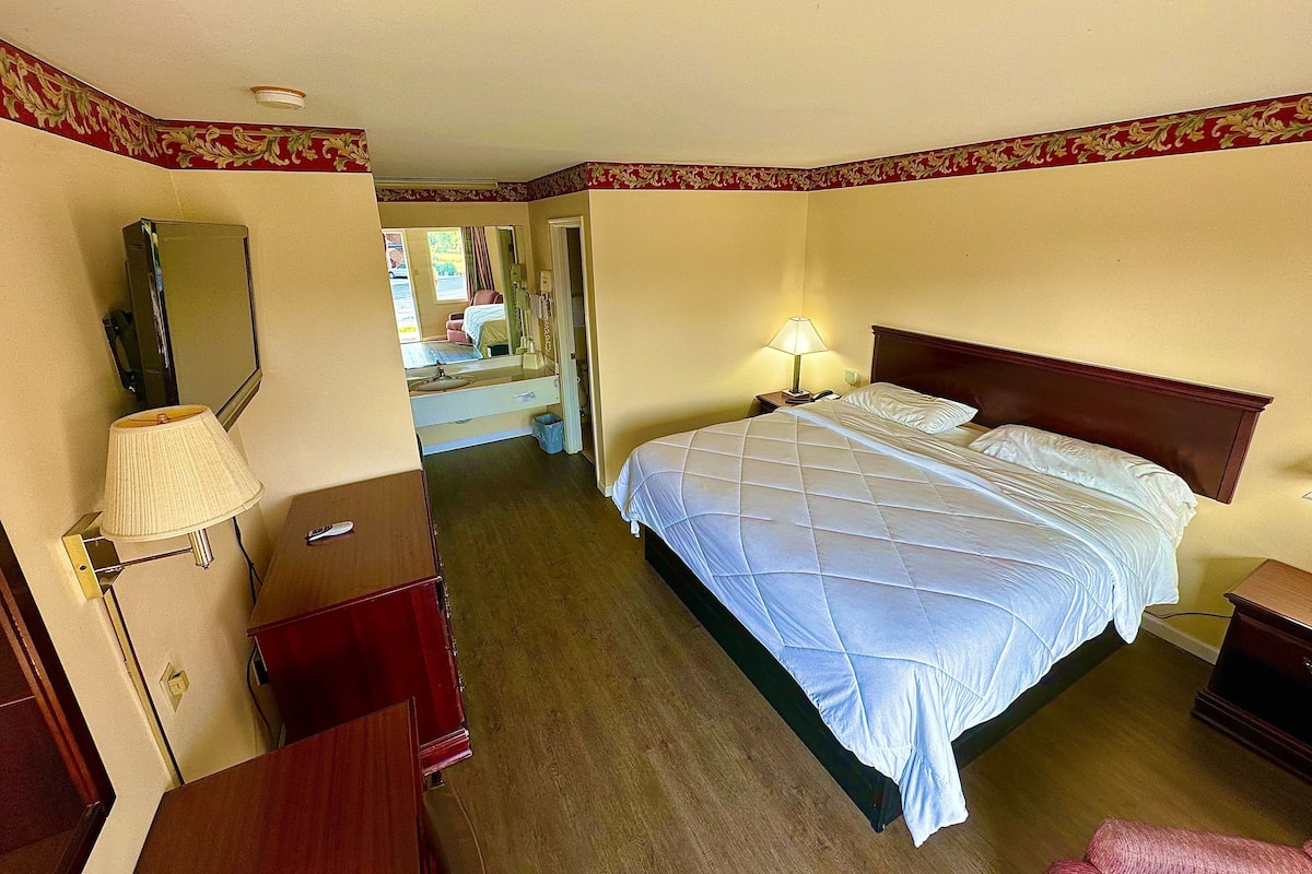 A hotel room with a bed, a desk, a mirror, a lamp, and a view of another room.