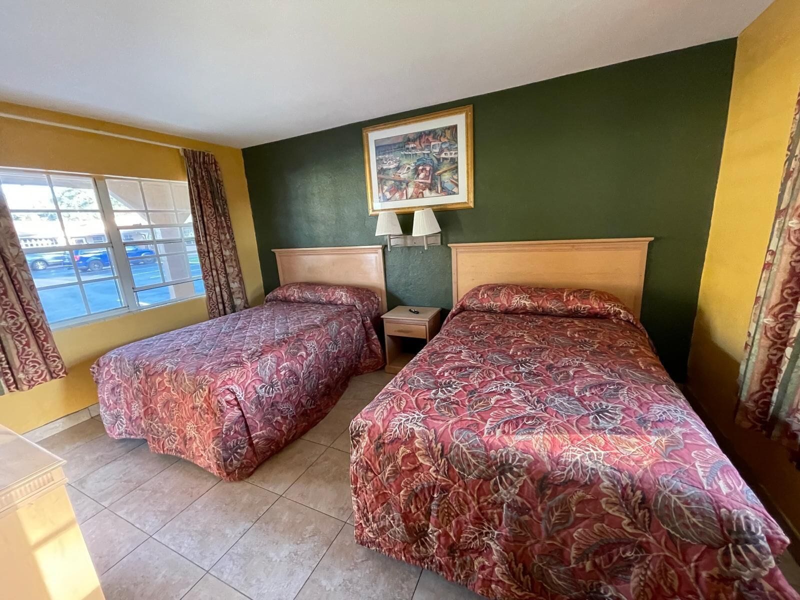 A hotel room with two beds, a nightstand, a lamp, a painting on the wall, and a window with curtains.