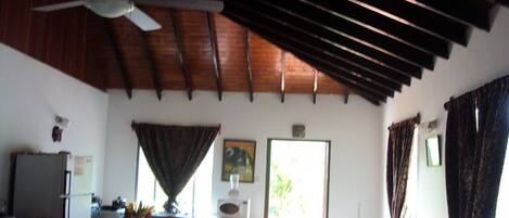 A spacious room with a wooden ceiling, a ceiling fan, a dining area with a table and chairs, and a kitchen area with a refrigerator and counter.