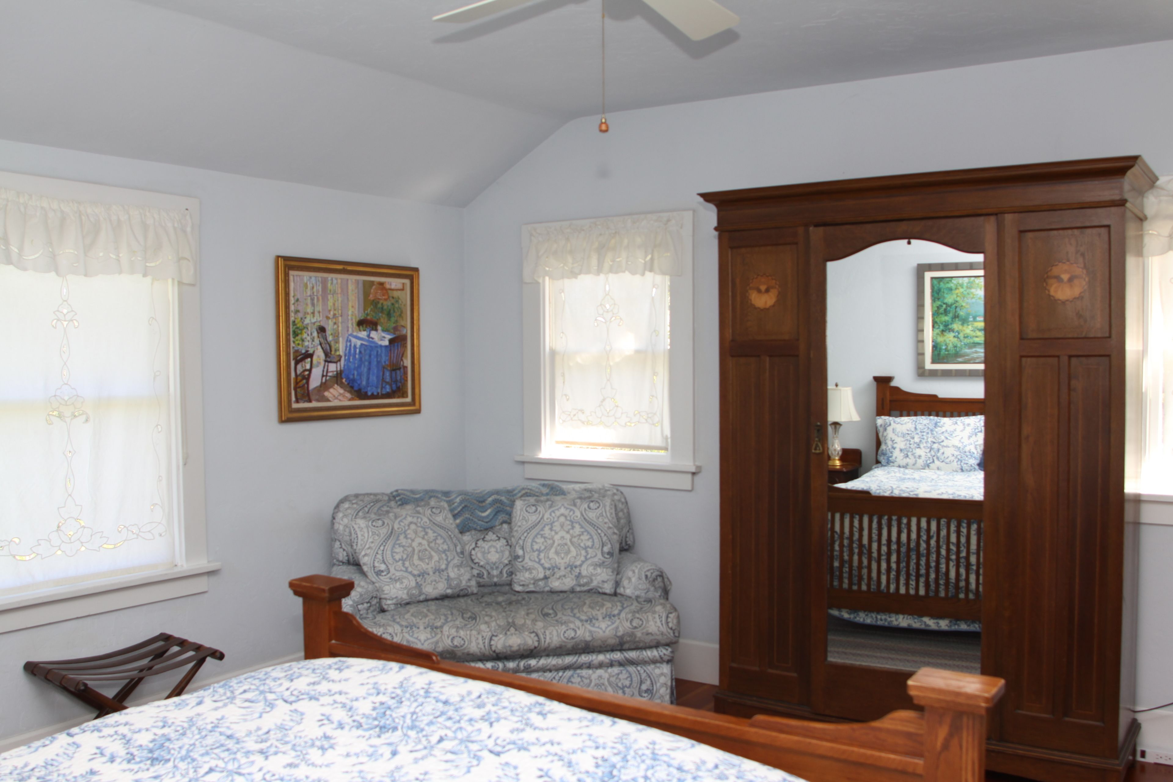 A bedroom with a bed, a wooden wardrobe, a patterned sofa, and two windows with curtains.