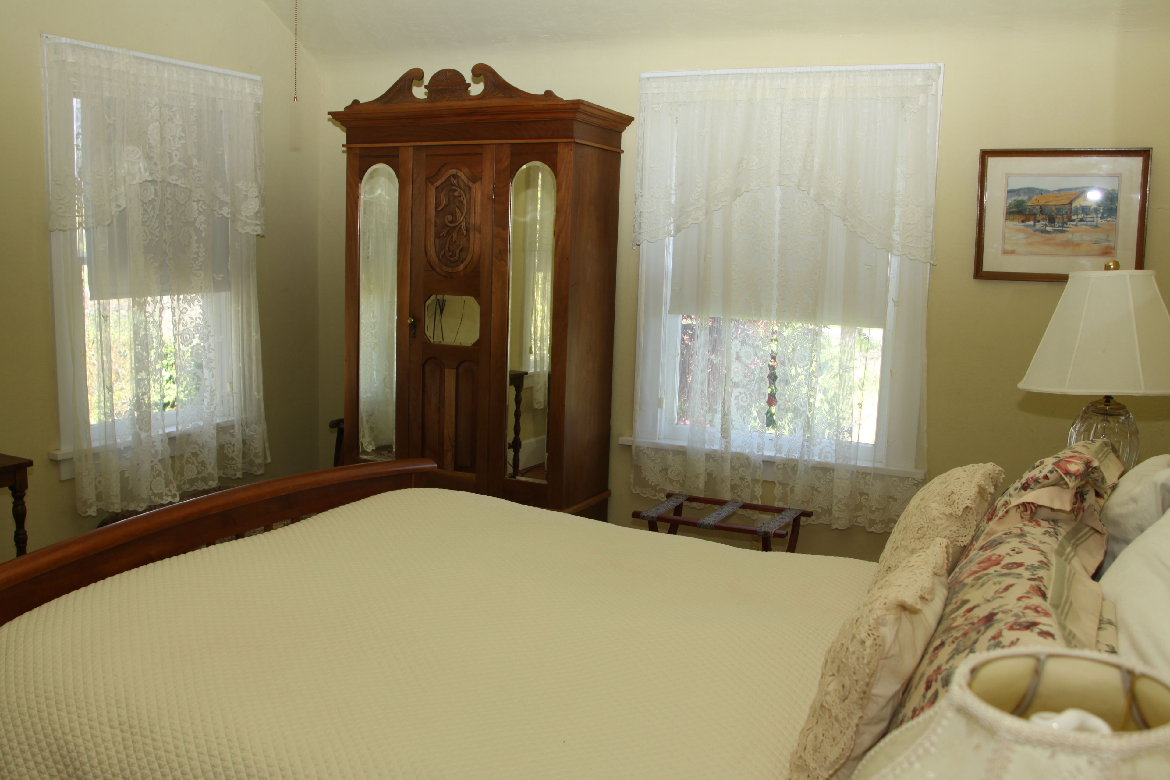 A bedroom with a large bed, a wooden wardrobe, lace curtains, a framed picture on the wall, and a bedside lamp.