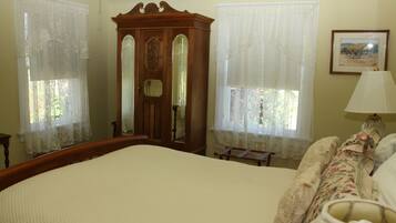 A bedroom with a large bed, a wooden wardrobe, lace curtains, a framed picture on the wall, and a bedside lamp.
