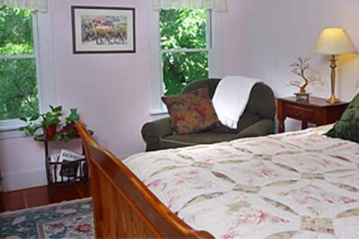 Une chambre à coucher comprenant un lit, une chaise, une table de chevet, une lampe et une fenêtre donnant sur un paysage verdoyant.