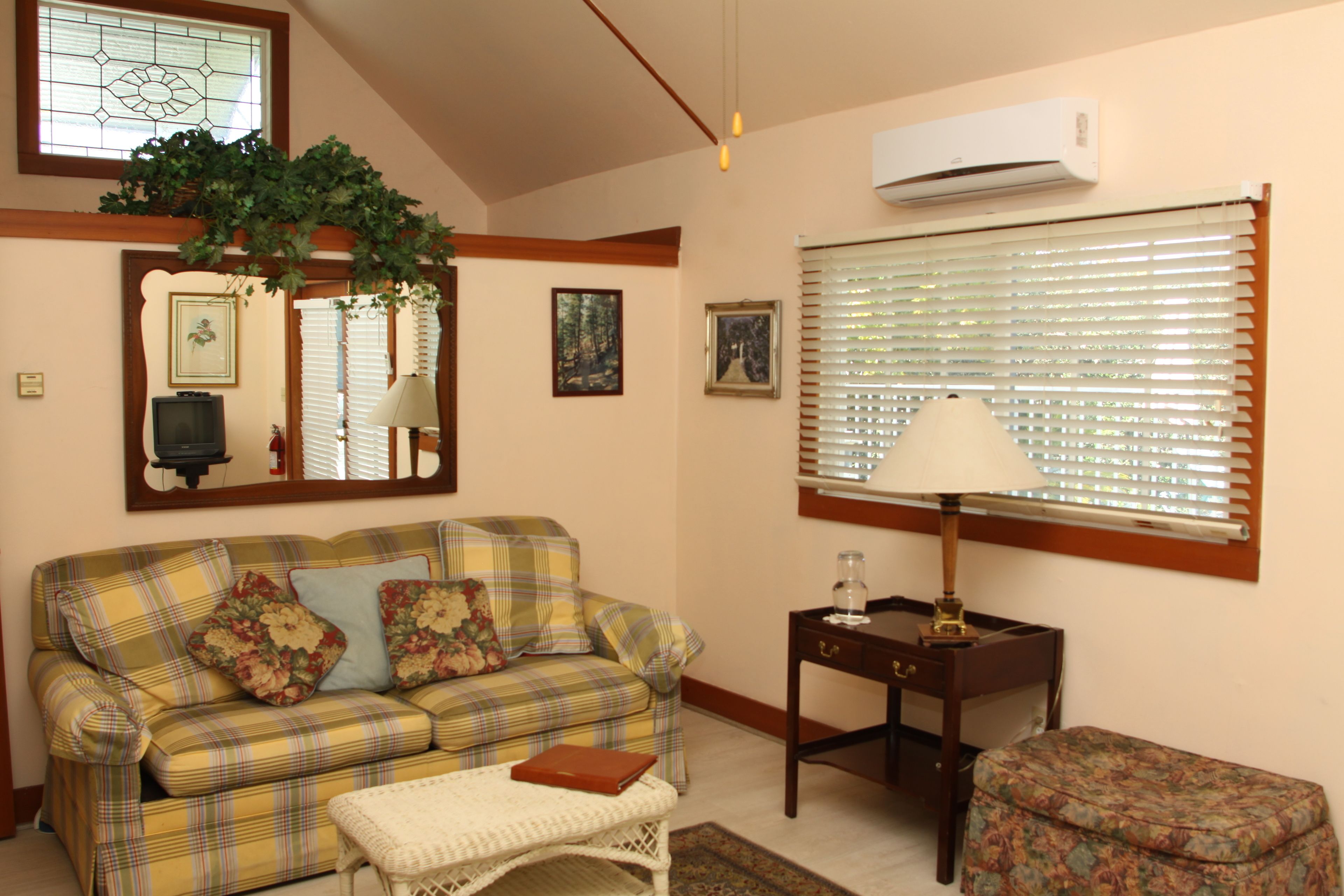 A cozy living room with a plaid sofa, a small table with a lamp, and a window with blinds.
