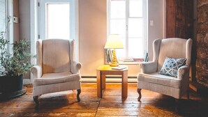 A room with two armchairs, a small wooden table with a lamp, and a window with shutters. - Le Clos des Brumes (Chateau-Richer)