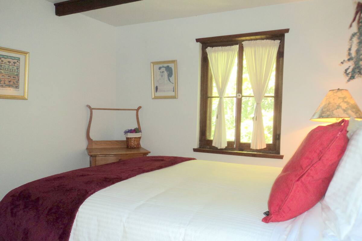 A bedroom with a bed, a window with curtains, a bedside table with a lamp, and a framed picture on the wall.