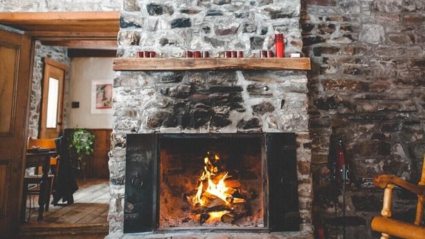 A stone fireplace with a fire, wooden shelves above, and a wooden railing on the left. - Le Clos des Brumes (Chateau-Richer)