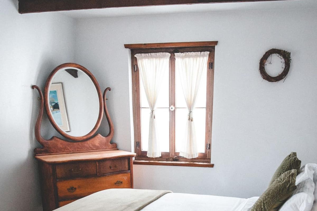 A bedroom with a bed, a dresser, a mirror, a window with curtains, and a wall clock.