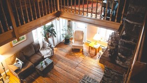 A spacious loft-style living area with a wooden floor, a sofa, a chair, and a table. There is a staircase leading to an upper level. - Le Clos des Brumes (Chateau-Richer)