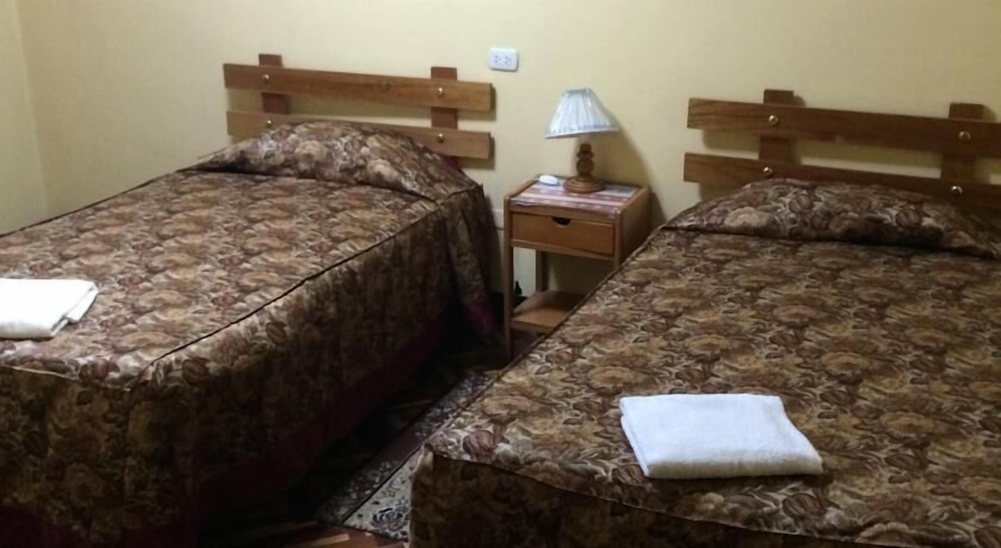 A room with two single beds, each with a wooden headboard and a patterned bedspread. There is a small bedside table with a lamp and a book on it.