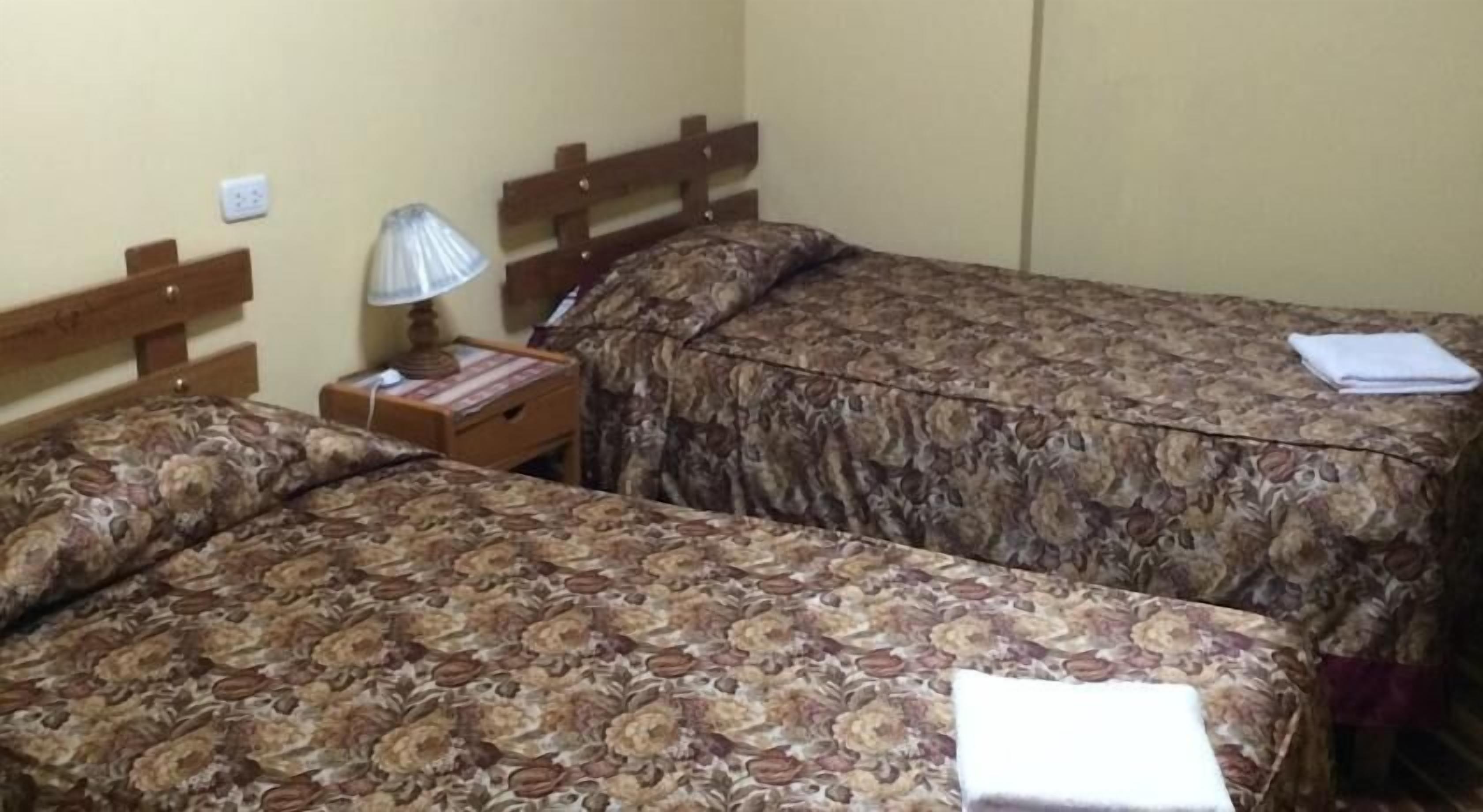 Una habitación con dos camas, cada una con colcha de estampado floral, una mesita de noche con lámpara y una toalla doblada en la cama derecha.