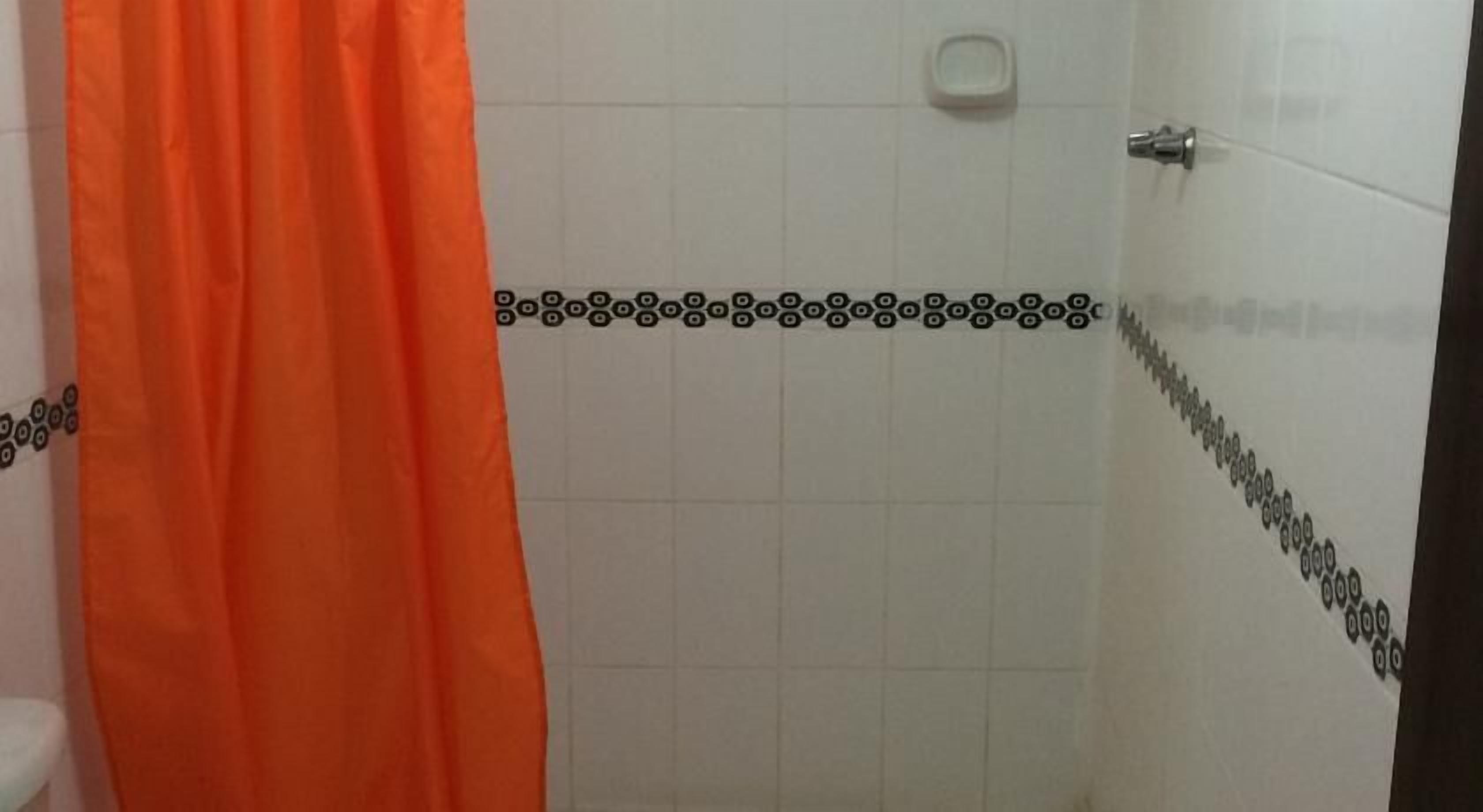Un baño con paredes de azulejos blancos, cortina de ducha naranja y un borde estampado en blanco y negro.