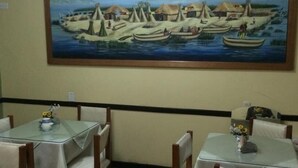 Una zona de menjador amb taules i cadires, un quadre d'un poble costaner a la paret i una decoració senzilla amb un vaset petit sobre la taula.