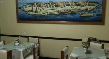 Un comedor con mesas y sillas, un cuadro de un pueblo costero en la pared, y una decoración sencilla con un pequeño jarrón sobre la mesa.