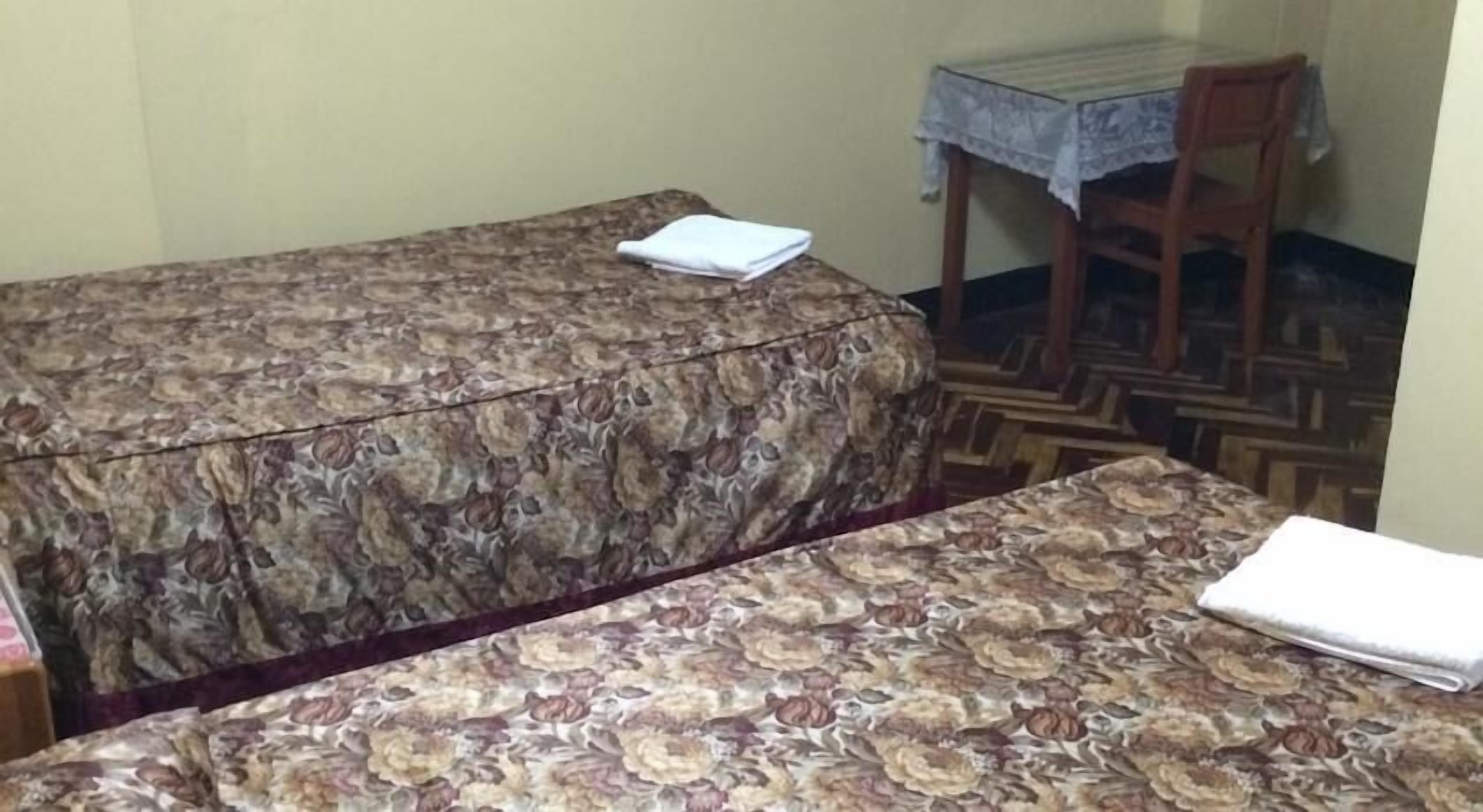 Um quarto pequeno com duas camas cobertas por roupa de cama estampada com flores, uma mesa de madeira com toalha de renda e uma cadeira.