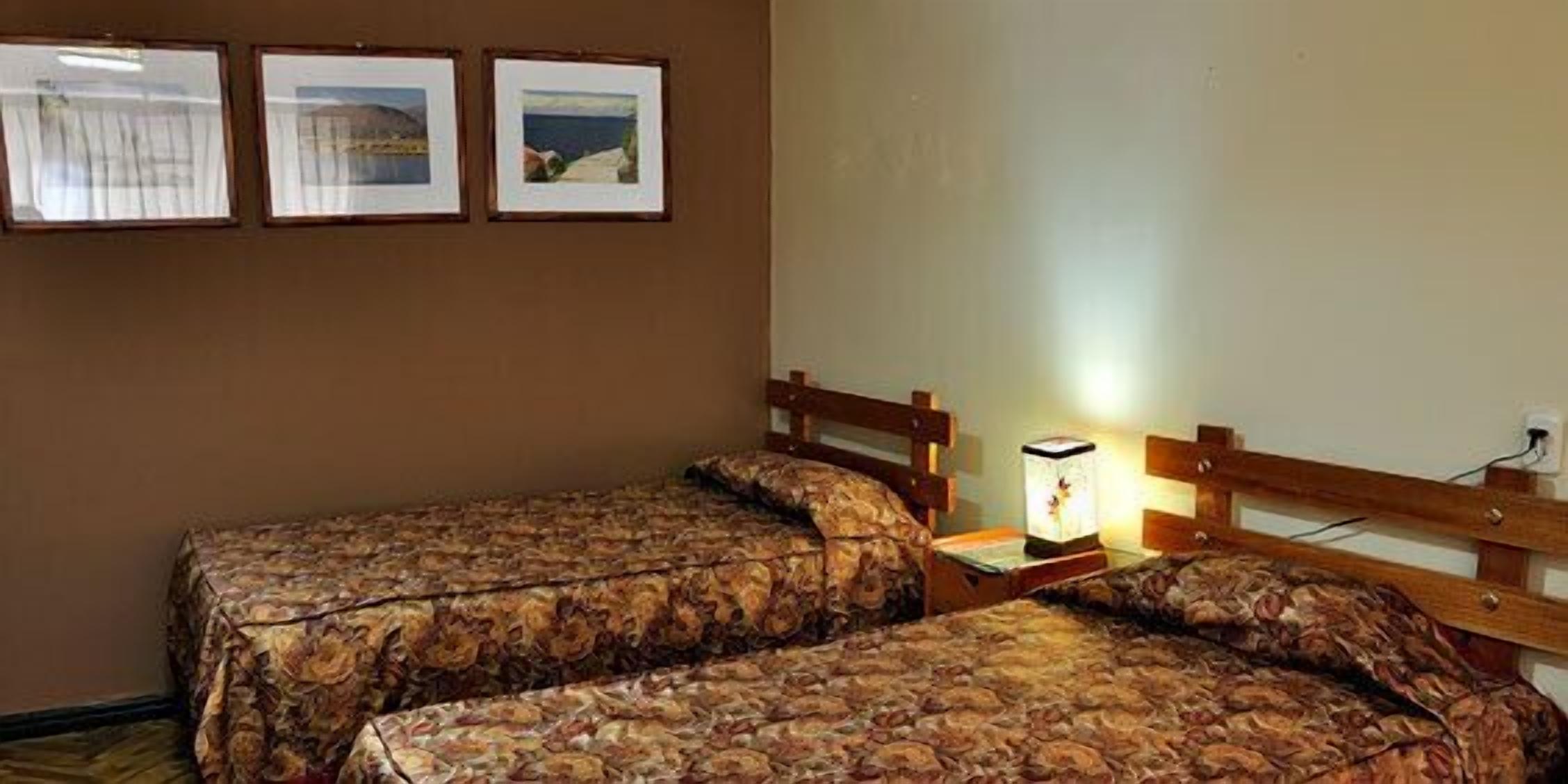 Una habitación con dos camas, una mesita de noche con lámpara y tres cuadros en la pared.