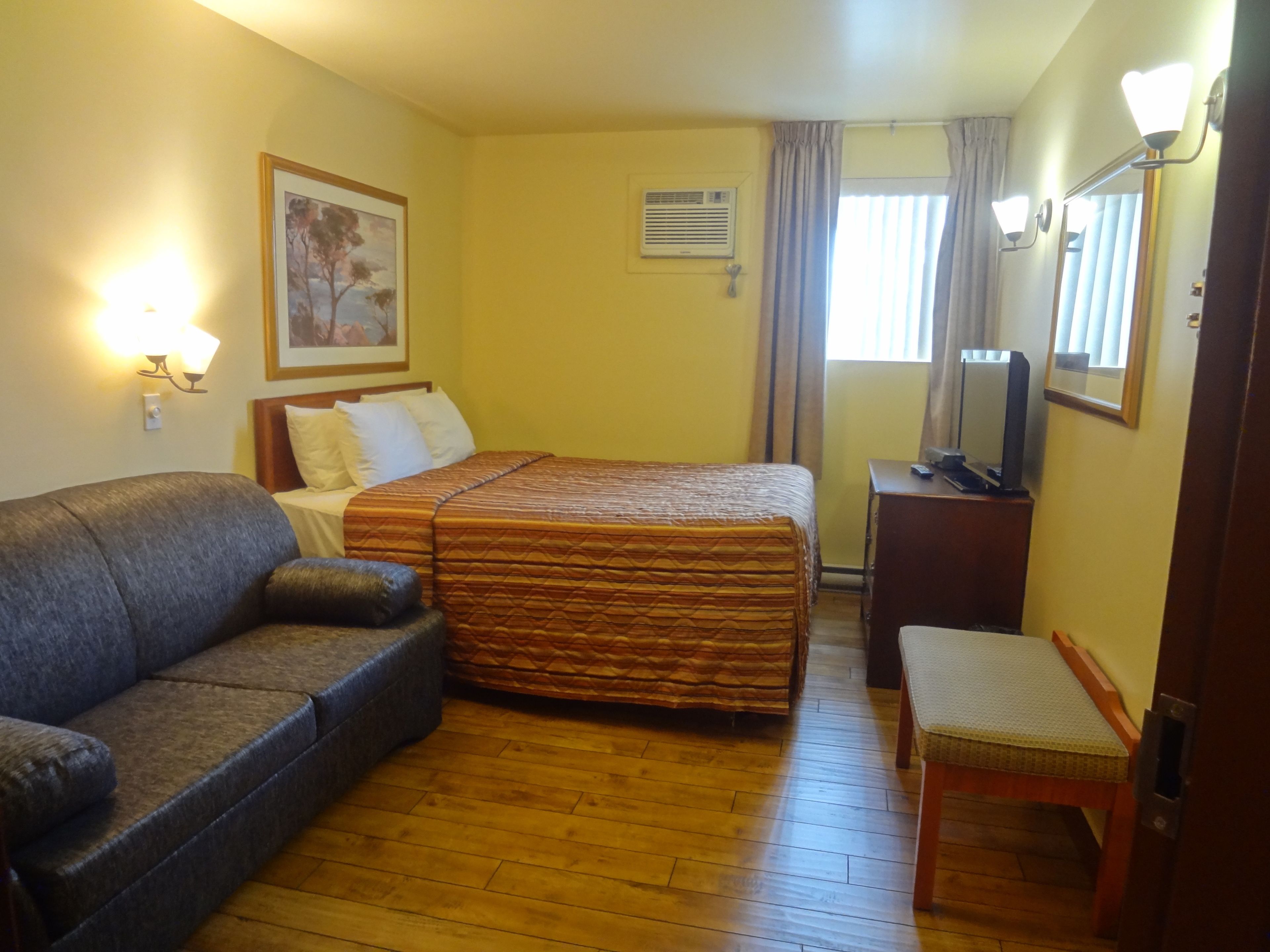 Habitación de hotel con cama, sofá, sillón, escritorio, espejo, cuadro y ventana con cortinas.