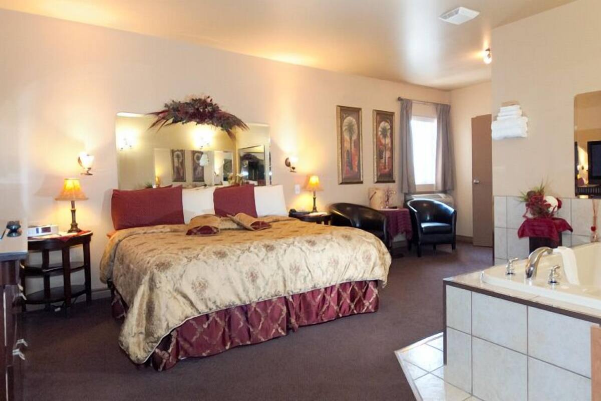 A hotel room with a large bed, a sitting area with a chair and a table, a television, and a bathtub with a red flower arrangement.
