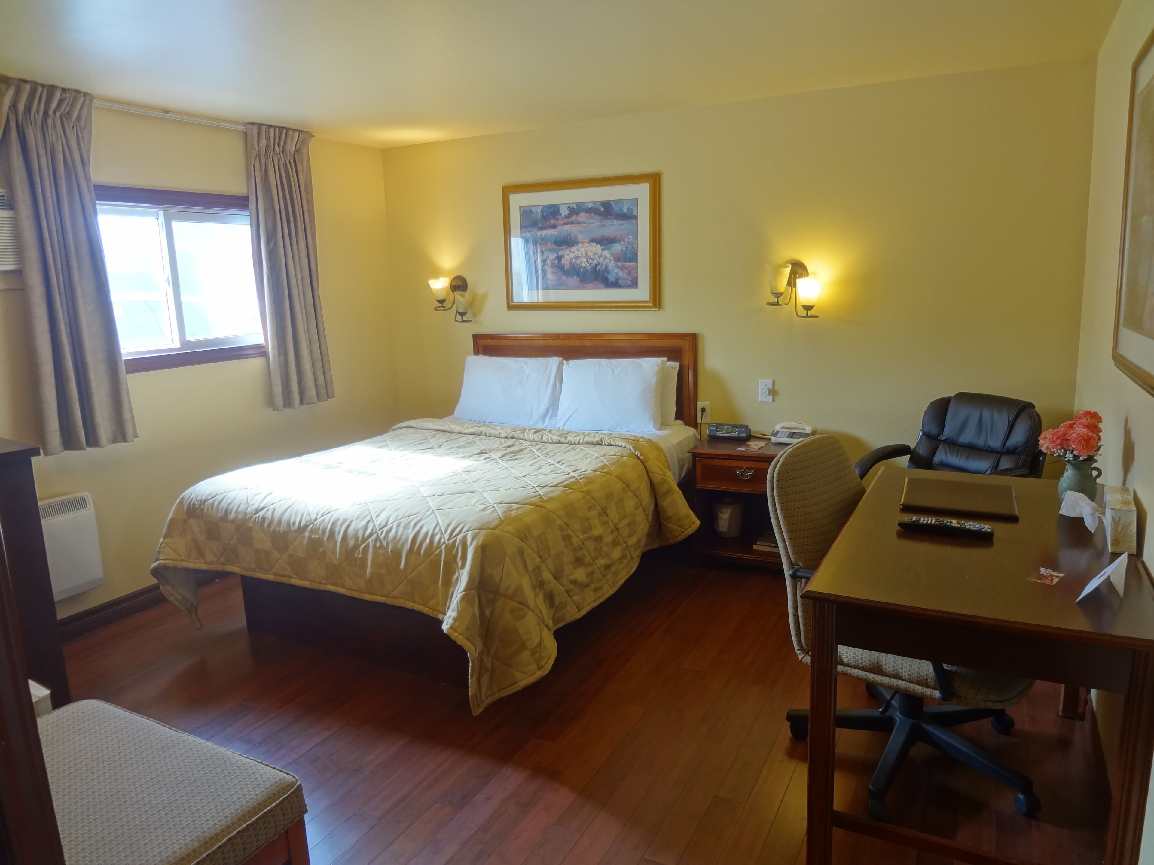 Una habitación de hotel con una cama, un escritorio con una silla, una lámpara, un cuadro en la pared y una ventana con cortinas.