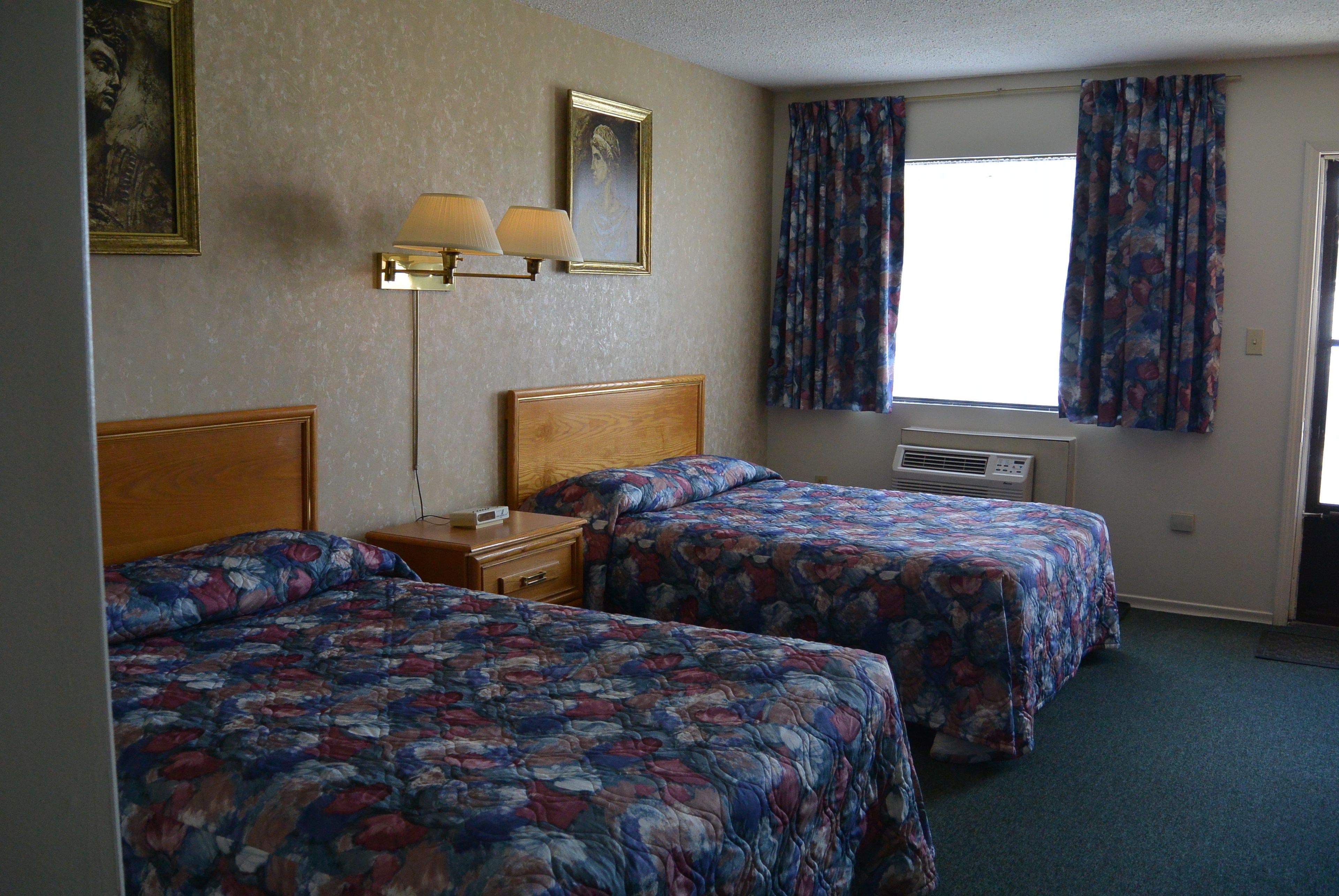 Hotelkamer met twee bedden, een nachtkastje, een lamp, een raam met gordijnen en een wandgemonteerde airconditioning.