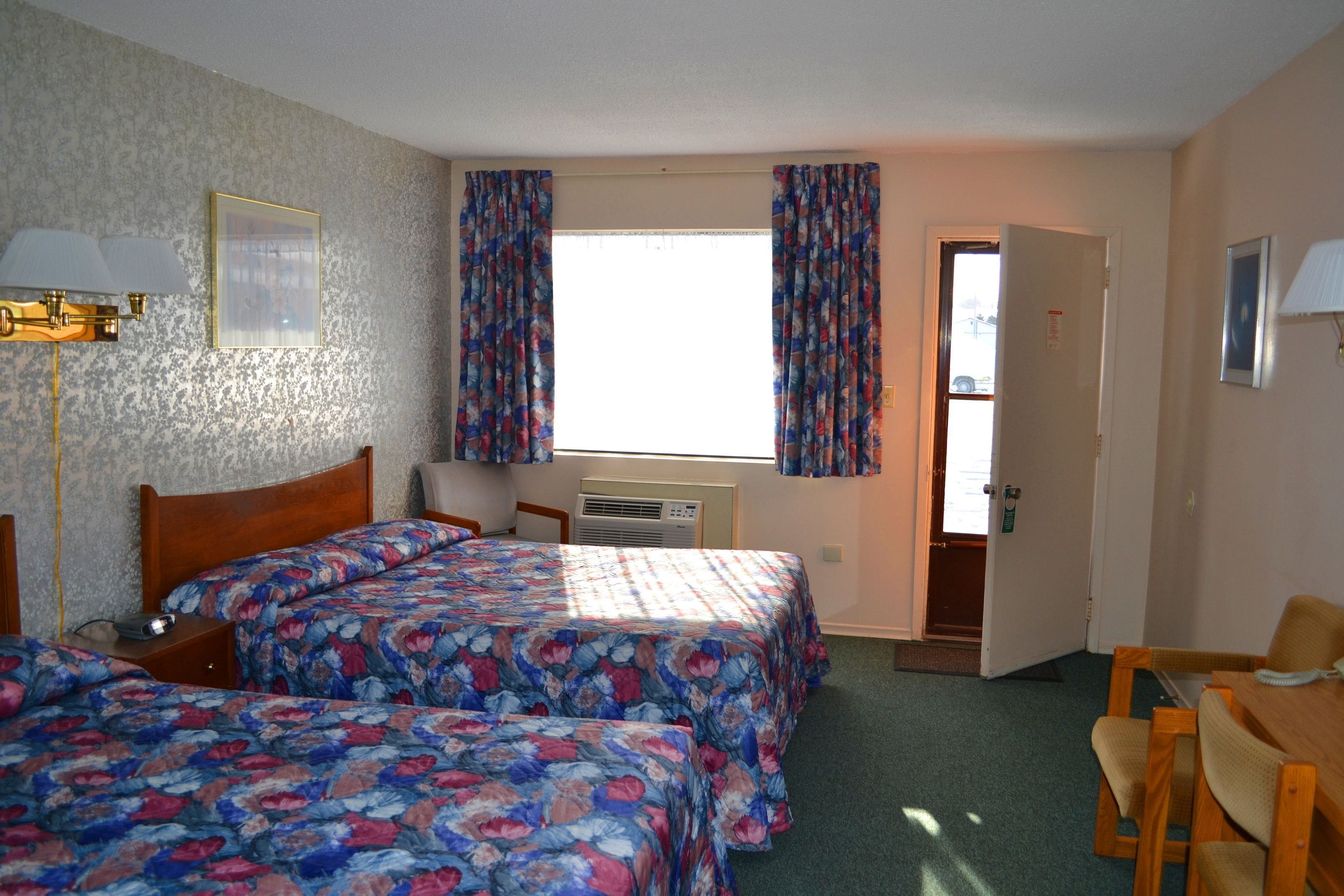 A room with two beds, a desk, and a chair. There is a window with curtains, a wall-mounted lamp, and a door leading to another room.