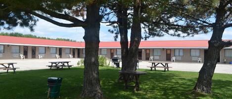 Un motel con techo rojo, mesas de picnic exteriores y grandes árboles en el jardín.