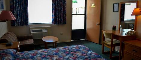 Habitación de hotel con cama, escritorio, silla, mesita, lámpara y ventana con cortinas.