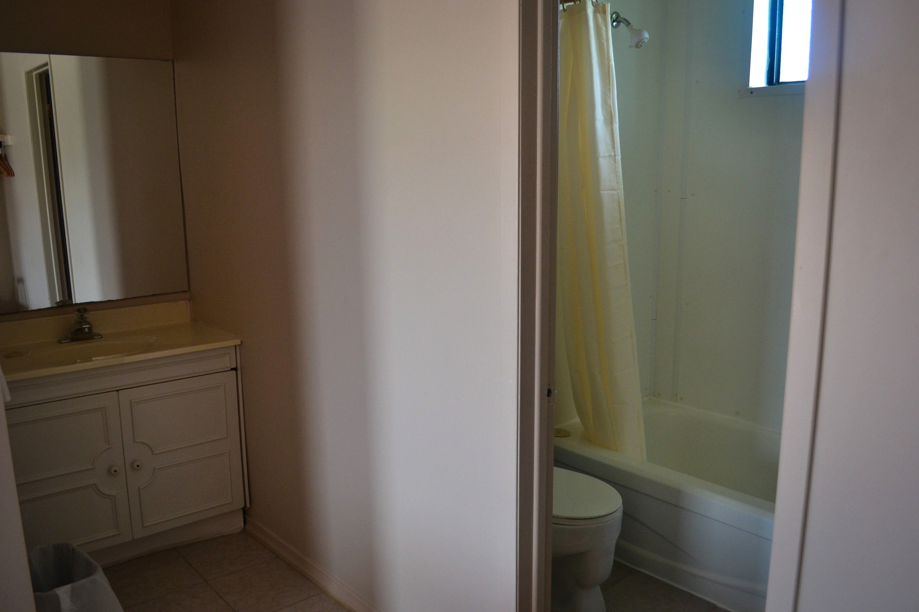 Une salle de bain comprenant des toilettes, une douche avec un rideau jaune, un lavabo avec un miroir et un meuble de rangement.