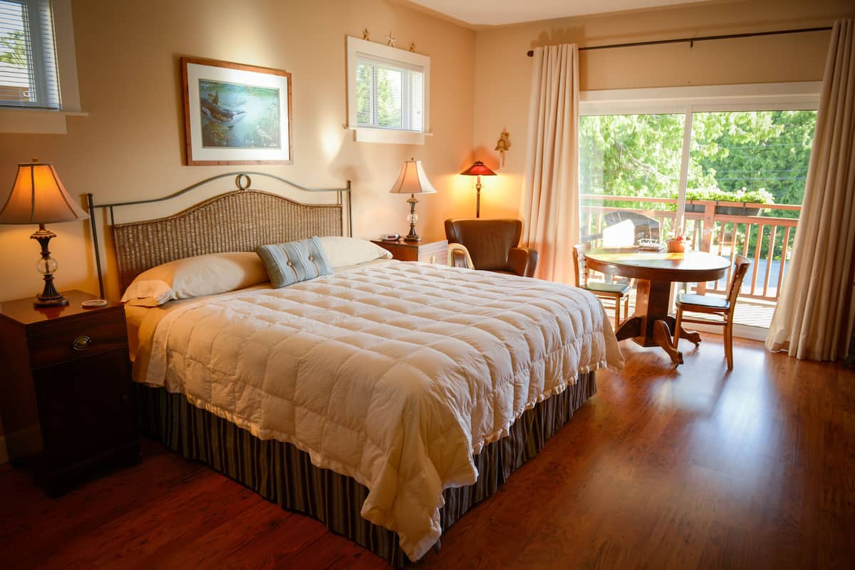 A bedroom with a large bed, a chair, a table, and a balcony with a view of trees.