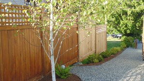 Un patio trasero residencial con cerco de madera, sendero de grava y un árbol pequeño.