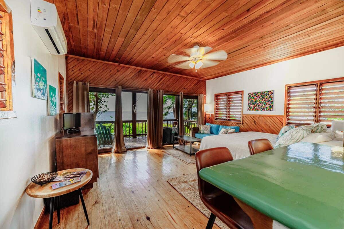 Amplia habitación con techo de madera, cama, escritorio y balcón con vista a palmeras.