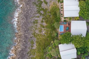 An aerial view of a coastal resort with a swimming pool, several white-roofed buildings, and lush greenery.