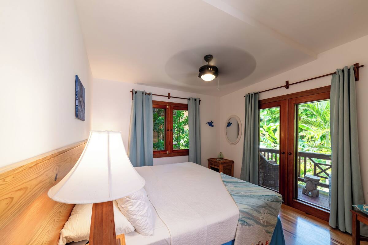 Un dormitorio con cama, mesita de noche, lámpara, ventana con cortinas y vista a zonas verdes.