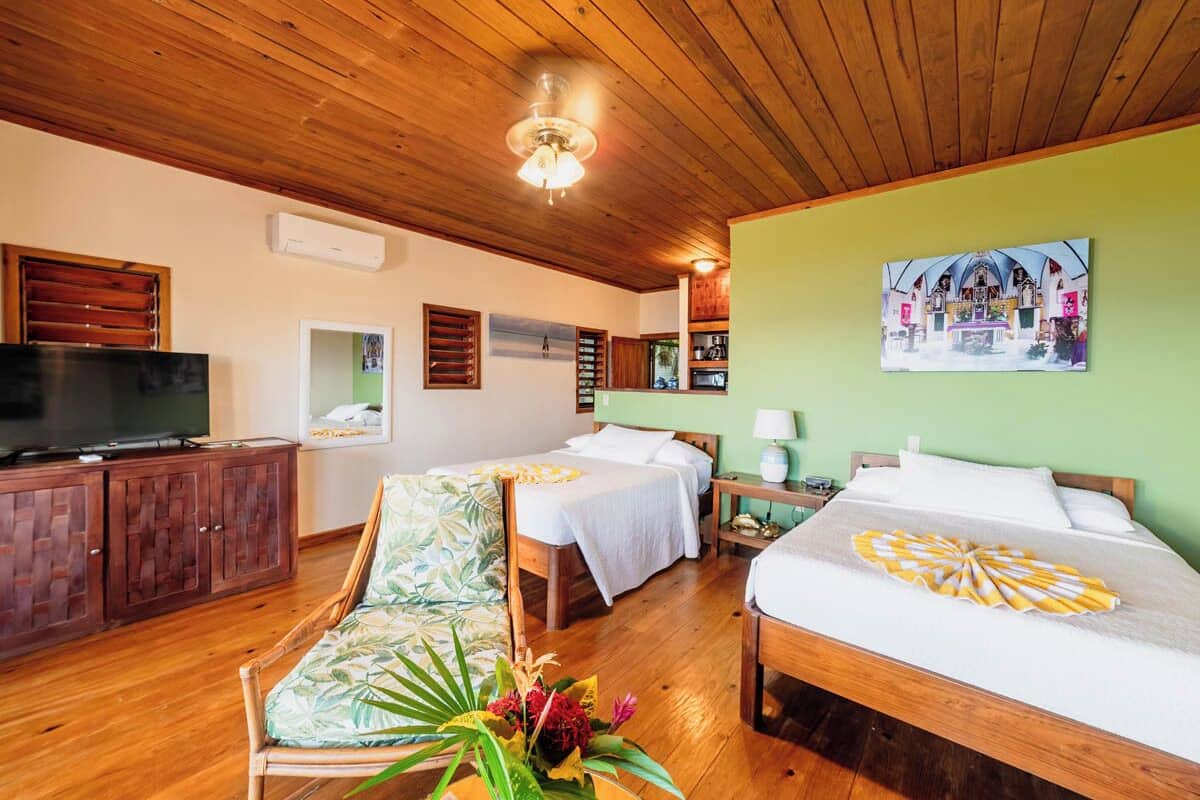 Habitación de hotel con piso de madera, televisor, dos camas, un sofá, ventilador de techo y un cuadro en la pared.