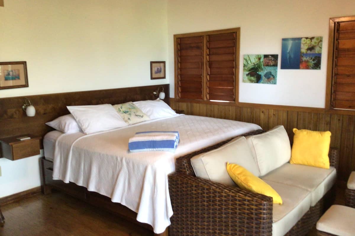 Un dormitorio con una cama, una mesita de noche, un sofá de mimbre con cojines y persianas de madera en las ventanas.