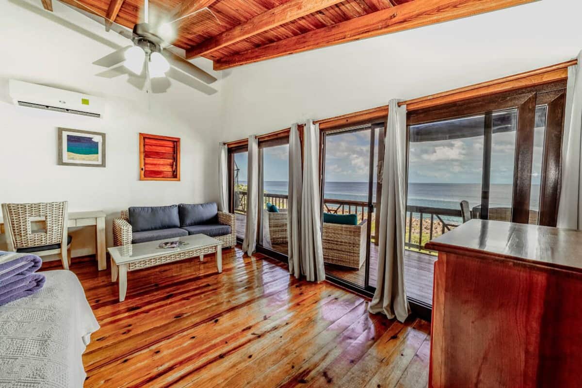 Una habitación con suelo de madera, ventilador de techo, sofá, mesa de centro, mobiliario de mimbre y ventanas amplias con vistas al océano.
