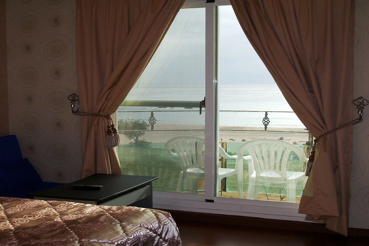 A bedroom with a bed, a nightstand, and a view of the beach through a sliding door.