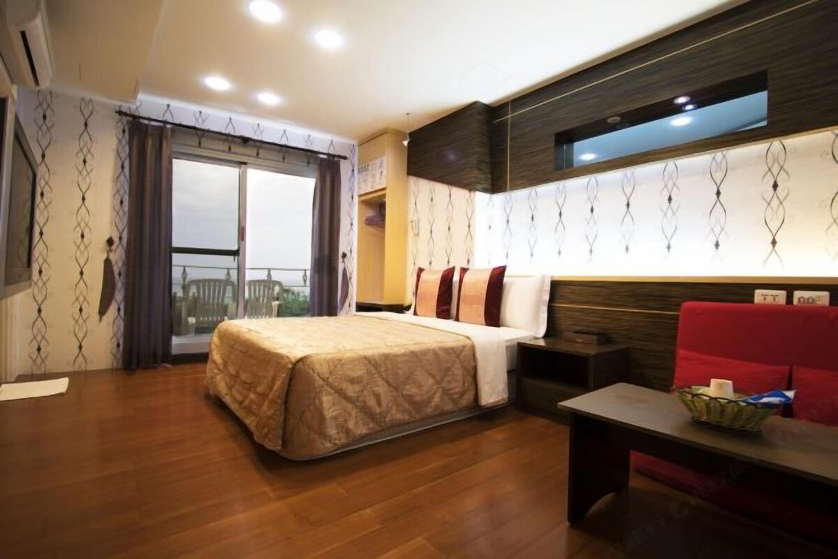 Habitación de hotel con cama, sofá rojo, una mesita y vistas al exterior a través de un ventanal grande.