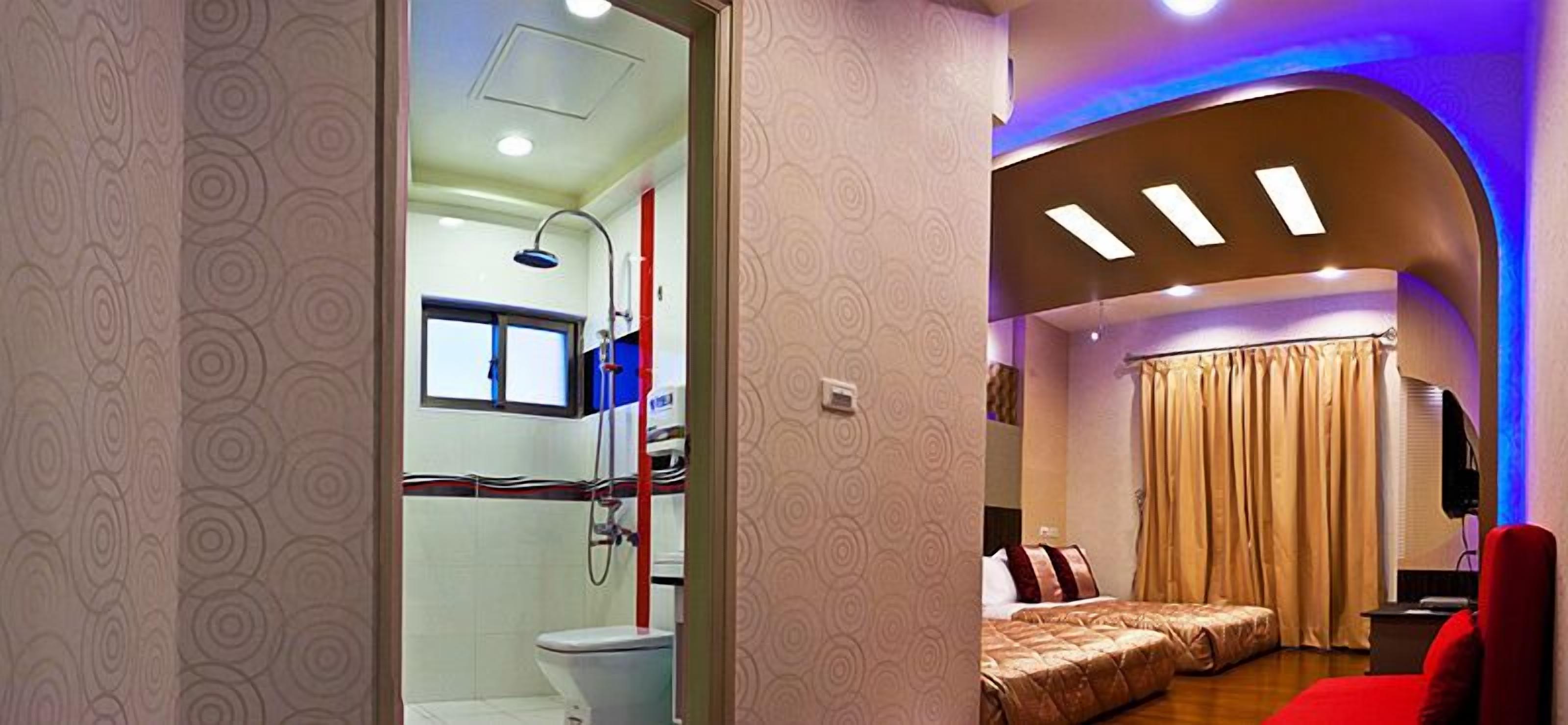 A modern bathroom with a shower, toilet, and a window. The room has a curved ceiling with recessed lighting and a bed with a red headboard in the adjacent bedroom.
