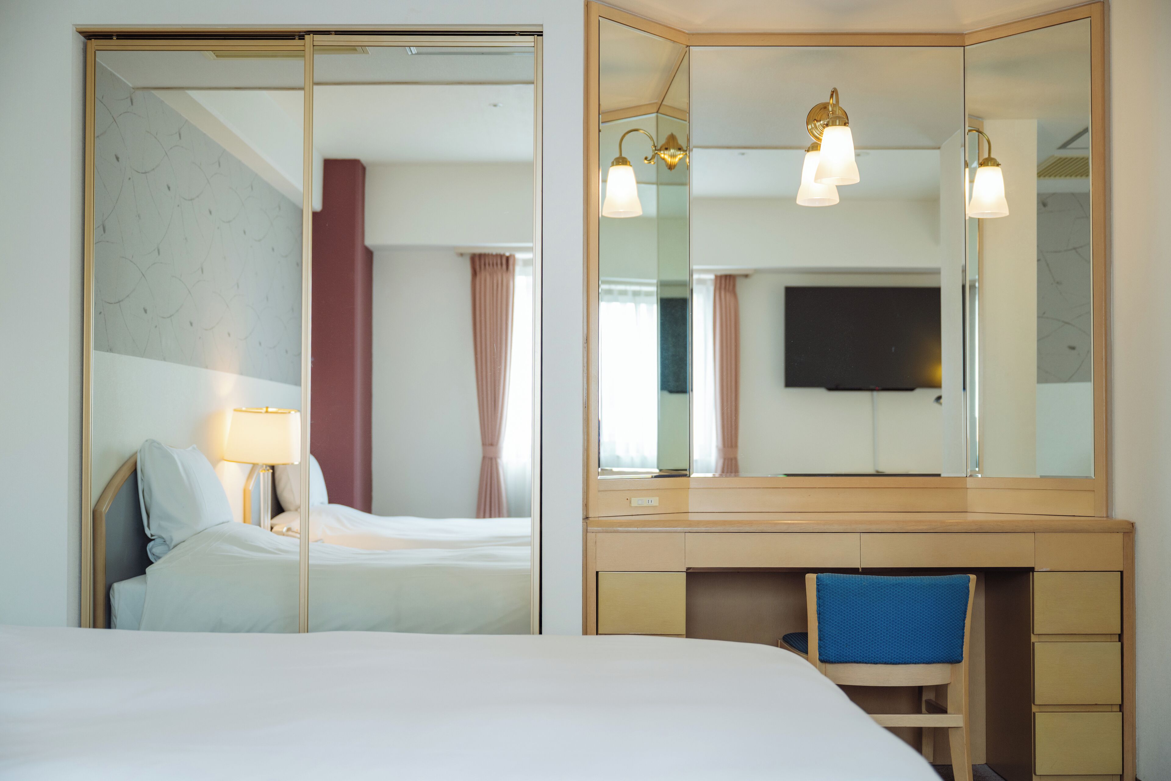 A hotel room with a bed, a desk with drawers, a chair, a TV, and a large mirror.